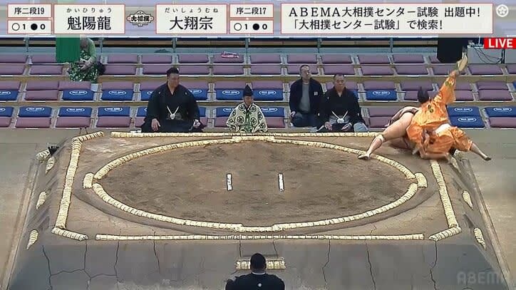 The wrestler fails to evade the approaching sumo wrestler... He falls out of the ring, but the professional spirit of the wrestler says, ``It's a match!''
